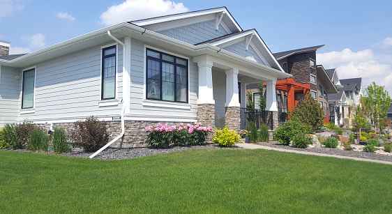 Calgary, Alberta, Canada- June 12,2023: New and modern homes including Craftsman style bungalow in foreground. Pink Peony in front along with shrubs and plants in a xeriscaped garden. Lawn in foreground.
