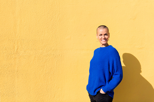 woman with a shaved head and a blue sweater smiling looking at camera with a yellow wall in the background, copy space for text