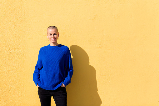 woman with a shaved head and a blue sweater smiling looking at camera with a yellow wall in the background, copy space for text