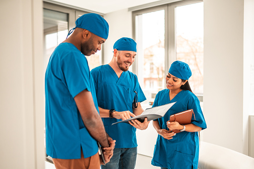Cheerful mid adult mixed race healthcare workers talking in a meeting at a modern medical center. They are in a meeting, talking and discussing a medical chart. They are wearing blue uniforms.