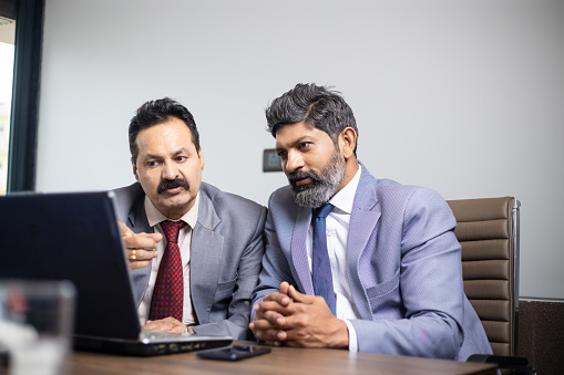 Two mature indian business men wearing suit working on laptop in office. Corporate people discussing work.