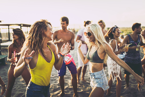 Crowd of young people having fun while dancing on a beach party in summer day. Focus is on blond woman dancing with her friend.