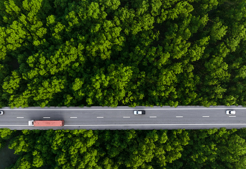 Aerial top view of car and truck driving on highway road in green forest. Sustainable transport. Drone view of hydrogen energy truck and electric vehicle driving on asphalt road through green forest.