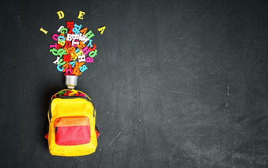 A light bulb symbol made from multicolored wooden letters, set by a vibrant backpack stuffed with the letters on a backboard background. The power of learning and imagination.