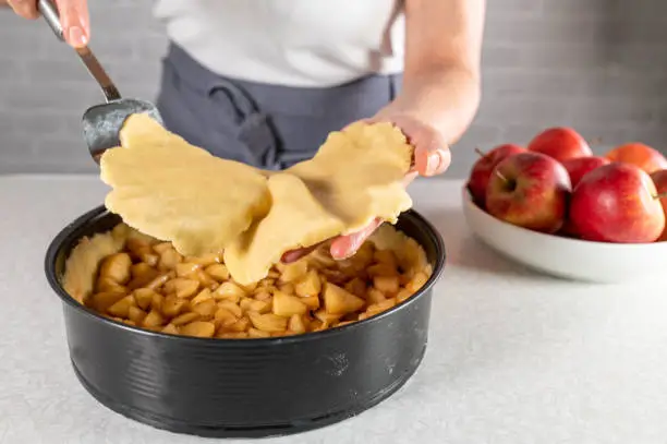 Womans hand ist putting a top crust or pie crust on top of a filled apple pie in a round baking pan. Traditional german "gedeckter Apfelkuchen".