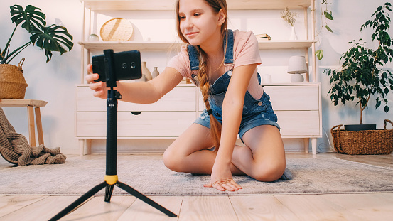 Kid vlog. Blogger home lifestyle. Create content. Happy smiling child girl adjusting phone camera on tripod recording video in light interior.