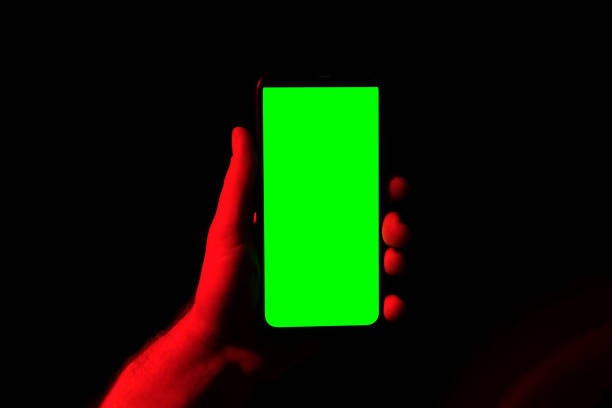 istock Mobile phone in young man hand. Use smartphone with green screen on red blue neon lighting, dark background. Male showing black phone chroma key display. Close up. Hand lifts device up holds it still 1498354606
