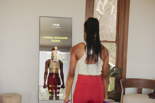 Young woman participating in a virtual workout class, using a smart fitness mirror to follow along with her instructor and get a comprehensive strength training workout in the comfort of her own home.