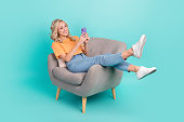 Portrait of cheerful nice person wear stylish t-shirt sit on armchair read email on smartphone isolated on turquoise color background