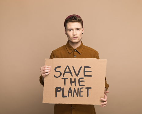 Portrait of young man wearing brown shirt and beanie holding „Save the planet” poster and looking at camera. Studio shot, brown background.