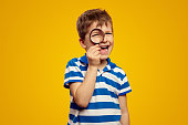 Curious schoolboy looking at camera through magnifier while standing against yellow background