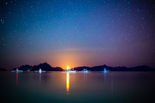 Night scene in El Nido, Philippines. The image features traditional Filipino boats peacefully floating on the sea. The sky above is adorned with bright stars, creating a stunning celestial backdrop. A prominent moon hangs above the horizon, casting a radiant glow on the surrounding landscape. The moon's intense light illuminates the silhouettes of distant islands, which are visible in the distance. The reflection of the moonlight creates a shimmering trail on the sea's surface. The color of the sky exhibits a gradient of blue and yellow due to the presence of the moon.