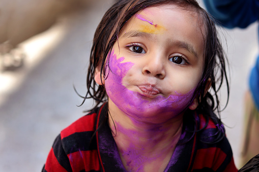 2-3 years baby girl with colored powder painted on both cheeks on Holi festival. Holi is a popular and significant Hindu festival celebrated as the Festival of Colours, Love and Spring. It celebrates the eternal and divine love of the god Radha and Krishna.