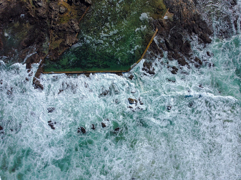 Aerial shot of a ocean pool on a rocky shoreline bashed by light blue and white ocean waves, Herolds bay.