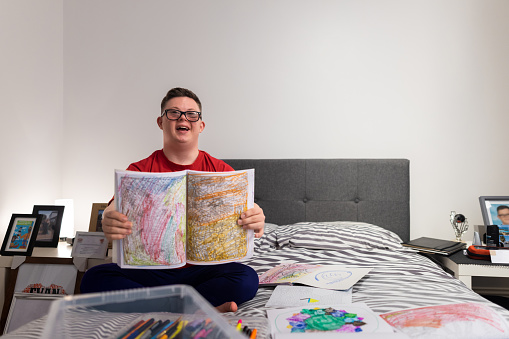 A full shot of a young male adult who has Down syndrome sitting on his bed surrounded by colouring pencils and colouring books. He is looking into the camera with positive emotion holding up a completed colouring book.
