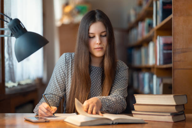 Smart young girl reading a book and taking notes, writing most important information she reads stock photo