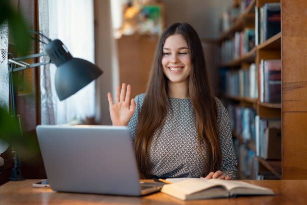 istock Diligent university student having online class from library, greetings with classmates to the webcam of her laptop 1498371062