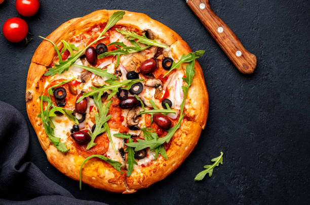 istock Hot pizza with spicy salami sausage, mozzarella cheese, mushrooms, black olives, tomato sauce and arugula, black table background, top view 1498373473
