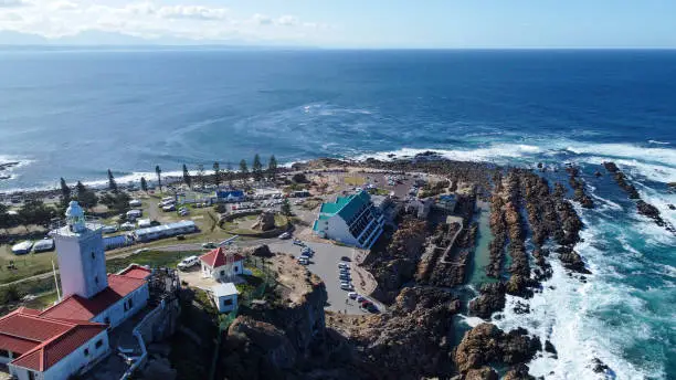 Aerial shot of the St Blaize lighthouse on top of a cliff looking down on the tip of Mossel bay with a view of various roads, buildings and a rocky shoreline bashed by white and blue ocean waves, Mossel bay.