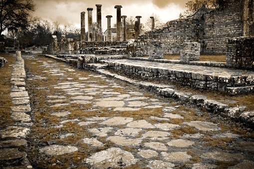 Ancient roman road in the archeological site of Saepinum, at Sepino, in the Molise region of Italy. In the background, the columns of a roman basilica. Sepia toned.