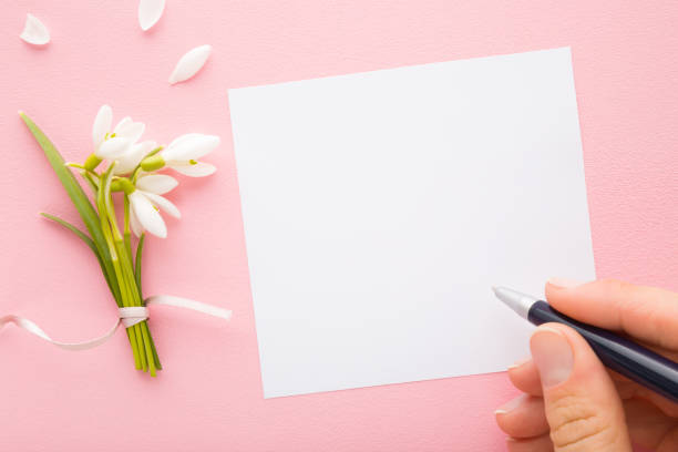 ペンを持ち、グリーティングカードに書く女性の指。明るいピンクのテーブルの背景に新鮮な白いスノードロップの束。パステルカラー。クローズ アップ。テキスト、引用、またはことわざ - invitation birthday card creativity ideas ストックフォトと画像