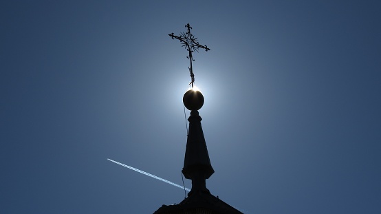 The blue sky with an airplane soaring against a tall church tower with a cross.