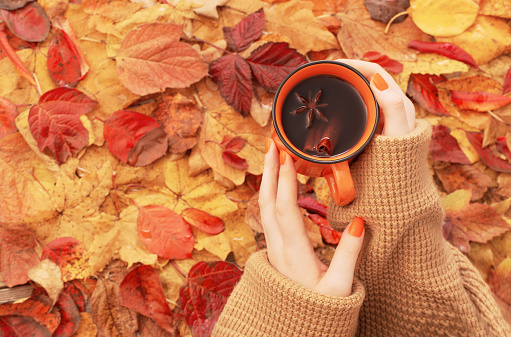 female hands with orange cup of coffee on background autumn leaves