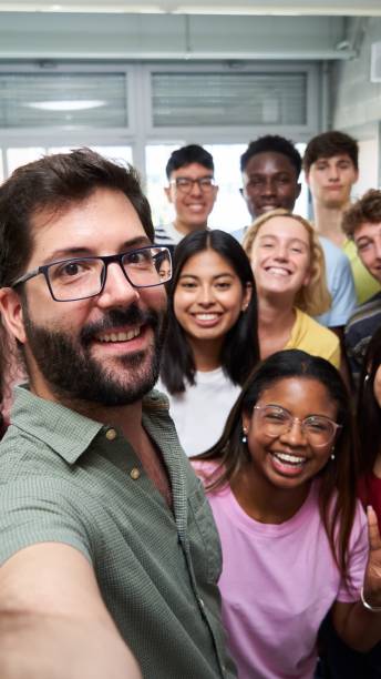 Happy selfie of young group of Erasmus students taking a photo with their teacher in the classroom. Happy vertical selfie of young group of students taking a photo with their male teacher in the classroom. Classmates from different countries, looking at camera with big smiles. mixed racial group of friendly children laughing together stock pictures, royalty-free photos & images