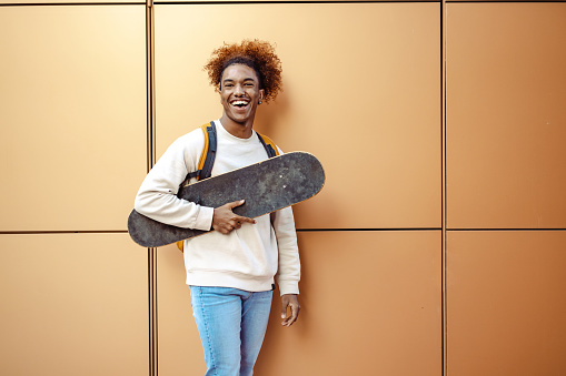 Urban young African American man holding a skateboard while standing on the street in front of the orange wall