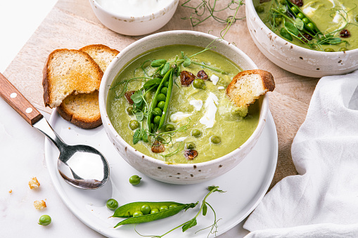 Two bowls with green pea cream soup with sour cream and toasts on white background.