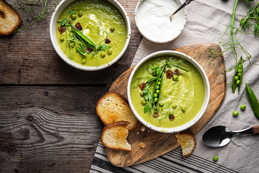 Two bowls with fresh green pea cream soup with sour cream and toasts on rustic wooden table.