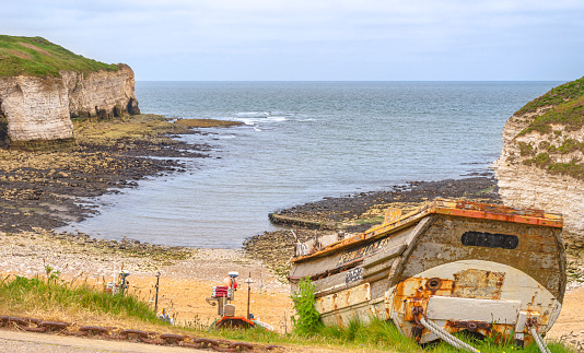 Flamborough, UK. June 9, 2023.  An old boat is the foreground pointing downwards to a beach with chalk cliffs on each side. A cloudy sky is above.