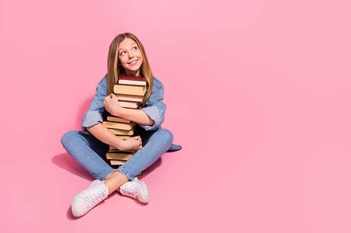 Photo of cheerful dreamy girl wear trendy jeans clothes sit floor hug hold many books look empty space isolated on pink color background.