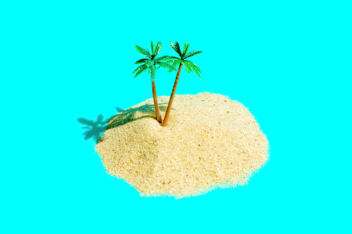 Remote tropical island composition made from sand pile and toy palm trees on a light blue background imitating the ocean. Exotic sandy beach summer vacation concept.