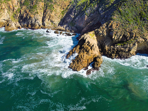 Aerial shot of a green coastal cliffside bashed by translucent light blue and white ocean waves, Herolds bay.