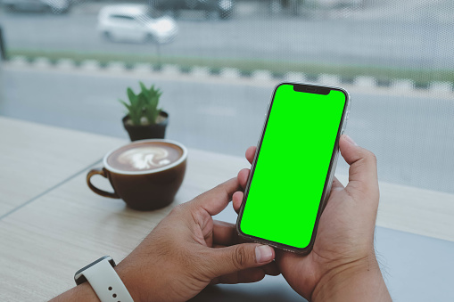 Mockup image of a business man holding smart mobile phone with blank green screen on vintage wooden table in modern cafe restaurant during meeting or lunch.