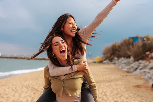 Happy young Caucasian woman carrying her Asian friend while walking down the beach on a rainy day.