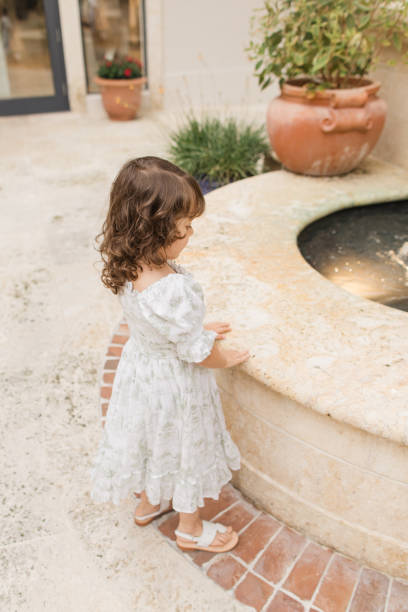 Lifestyle Candid Moment of a Cuban-American 4-Year-Old Toddler Girl with Brown Curly Hair Wearing Sage Green & White Spring Smocked Dress, a Hair Bow & White Leather Sandals While Walking in a Tuscan-Inspired Garden in Palm Beach, Florida in May of 2023 momento sincero de estilo de vida de una niña cubana-americana de 4 años con cabello castaño rizado con vestido de bata de primavera verde y blanco, un lazo para el cabello y sandalias de cuero blanco mientras camina en un jardín de inspiración toscan