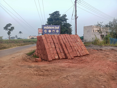 Kolhapur, India- November 1 st 2022; stock photo was captured under natural light, depicting a pile of red bricks positioned near the roadside, with a blue-colored board placed adjacent to the bricks.