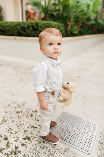 Lifestyle Candid Moment of a Cuban-American 1-Year-Old Baby Boy with Short Light Brown Hair Wearing Sage Green Bow Tie, Suspenders, White Buttondown Shirt, Pants & Brown Shoes Walking Around a Garden in Tropical Palm Beach, Florida in May of 2023