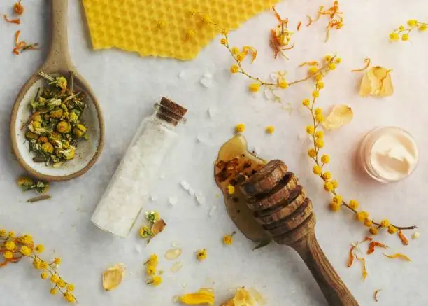 An assortment of homemade body care products and vibrant flowers arranged on a white surface with a wooden spoon beside it