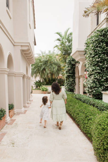Lifestyle Candid Moment of a Cuban-American 4-Year-Old Toddler Girl with Her 31-Year-Old Cuban Mother Both w/ Brown Curly Hair Wearing Sage Green & White Spring Smocked Dress & White Leather Sandals While in a Tuscan-Inspired Garden in Palm Beach, Florida niña cubana estadounidense de 4 años caminando mientras se toma de la mano con su madre cubana de 31 años con cabello castaño rizado con vestido de primavera verde y blanco salvia y sandalias de cuero blanco mientras está en un jardín inspirado en la