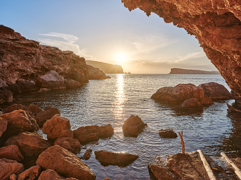 Wide-angle view of one of the most scenic and secluded coves of Platges de Comte (also known as Cala Conta), on the western coast of Ibiza, characterised by rough, shallow cliffs and the presence of a wooden mooring pole and slipway for boats. Blue sky, picturesque clouds, calm waters reflecting the oblique, golden light of a Mediterranean summer sunset, the gentle splashing of the waves on the shore. The islets of S'Illa des Bosc and Sa Conillera, with their peculiar outlines, occupy the background. High level of detail, natural rendition, realistic feel. Developed from RAW.