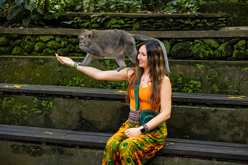 A young woman is sitting on a wooden bench as a female macaque balances on her outstretched arm. The macaque is eating corn from her hand.