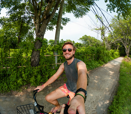 Young man taking a selfie while cycling around the island of Gili Air. He's wearing swimming shorts, a vest and sunglasses. He is looking at the camera and smiling.