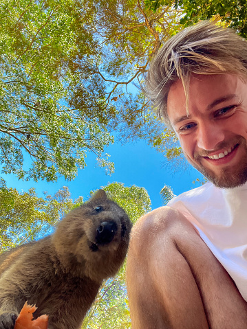 Low angle view of a young man taking a selfie with a Quokka. They are both looking down to the camera and smiling.
