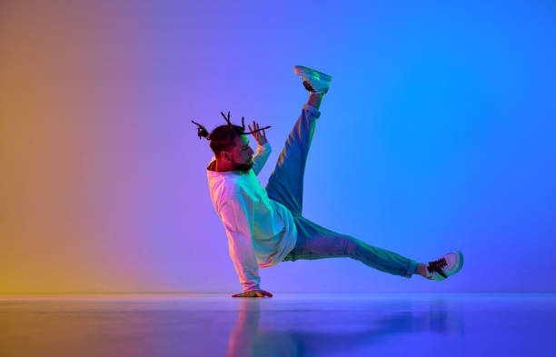 Young handsome guy in casual clothes and dreads dancing breakdance against gradient multicolored studio background in neon light