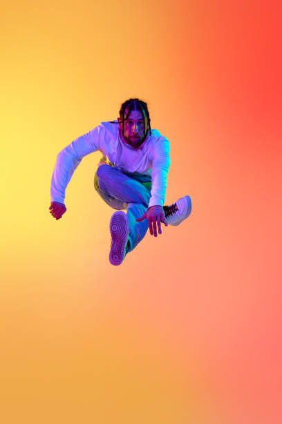 istock Young bearded man with dreads in casual clothes dancing breakdance against gradient orange yellow studio background in neon light 1498420768