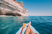 Men's legs and paddle on a supboard against the backdrop of a picturesque seascape with a rock. Relaxation and fitness on the water