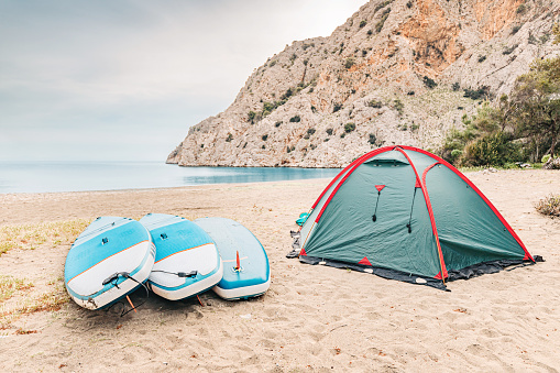Camping tent with sup boards on the wild beach at mediterranean seacoast. Water trip and sportive recreation